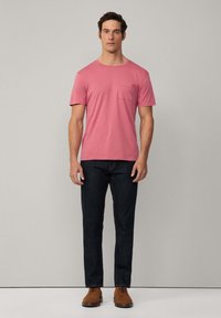 Muška kratka majica ružičaste boje s džepom na prsima, u kombinaciji s tamnim trapericama i smeškim čizmama sa vezicama, na neutralnoj pozadini.