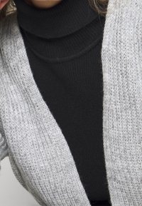 Osoba w czarnym prążkowanym golfie pod jasnoszarym dzianinowym kardiganem, pokazująca górną część tułowia i szyi.