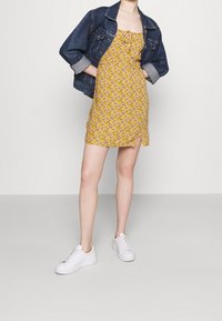 Gul blommig klänning med en åtsittande bodice, knälång och med en sidoklyvning, lager under en jeansjacka. Matchas med vita sneakers.