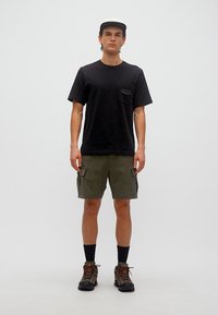 Νεαρός άνδρας που φοράει μαύρο καπέλο, μαύρο μπλουζάκι, χακί βερμούδες cargo, μαύρες κάλτσες και παπούτσια πεζοπορίας, στέκεται μπροστά σε απλό φόντο.