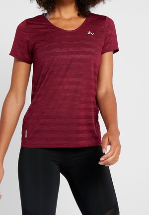 Vrouw die een nauwsluitend bordeauxrood sportshirt met korte mouwen en zwarte legging draagt, poseert met ontspannen armen tegen een effen achtergrond.