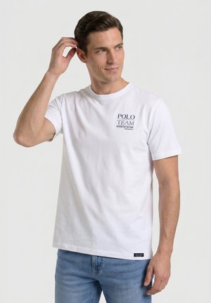 Hombre con cabello oscuro que lleva una camiseta blanca Polo Team y vaqueros azules, tocándose la cabeza y mirando ligeramente hacia la derecha contra un fondo liso.