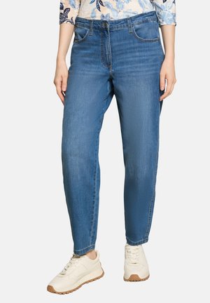 Kvinde iført blå denimjeans, blomstret top og hvide sneakers står med hænderne afslappet ned langs siderne.
