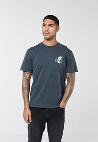 Muškarac s tetovažama na oba nadlaktica, nosi tamnoplavu majicu s bijelim grbom orla i zvijezda, stoji s jednom rukom u džepu.