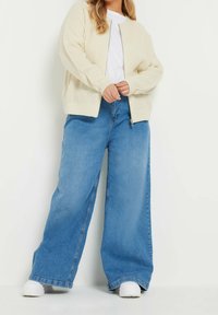 Person som bär en krämfärgad stickad zip-up cardigan, vit T-shirt, vida blå jeans och vita sneakers står mot en vit bakgrund.