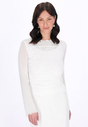 Femeie cu păr ondulat închis poartă cercei de perle tip picătură și un top alb mulat cu mâneci lungi din material transparent, pe un fundal simplu.