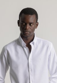 Hombre de piel oscura vestido con una camisa blanca de botones, mirando hacia su izquierda contra un fondo liso de color gris claro.
