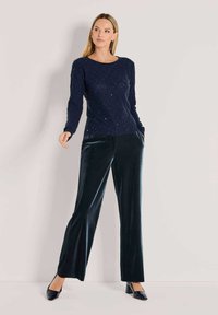 Frau steht mit einem marineblauen, gesprenkelten Pullover, dunklen, weiten Samenhosen und schwarzen Blockabsatzschuhen vor einem einfarbigen Hintergrund.