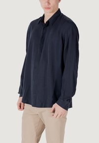 Navyblauwe langzamige shirt van gladde stof, met een klassieke kraag, sluiting met knopen aan de voorkant, en knoopsluiting bij de mouwen, gedragen met beige broek.