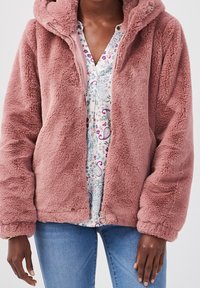 Person iført en lynlås dusty pink, blød hættejakke over en hvid bluse med paisley-mønster og blå jeans, stående med afslappede arme.