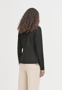 Veste noire sur mesure à manches longues, présentant une silhouette ajustée et un tissu lisse. Portée avec un pantalon beige clair, vue de dos.
