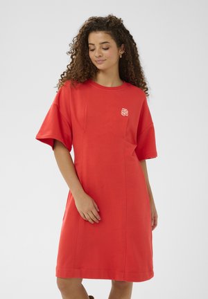 Femme aux cheveux bouclés portant une robe rouge ample, longueur genou, à manches courtes, avec un petit logo blanc sur la poitrine, sur un fond uni.