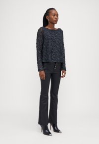 Zwart doorschijnend langemouwen top met textuur borduursel, gecombineerd met fitted zwarte broek met hoge taille en knoopdetails, en puntige enkellaarzen.