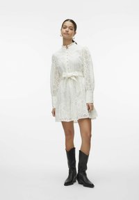 Abito bianco in cotone con ricami, orlo a volant, maniche lunghe, chiusura a bottoni e cintura da annodare in vita. Indossato con stivali da cowboy neri. Tessuto morbido.