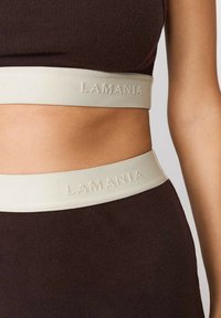Hnědý pružný sportovní top s jemně béžovým elastickým pásem s vyšitým nápisem "LAMANIA". Detailní pohled na texturu a barevný kontrast.