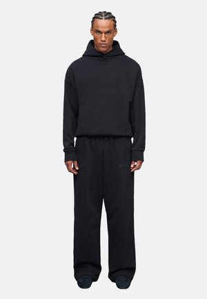 Sudadera negra de gran tamaño con un bolsillo canguro, combinada con pantalones de chándal negros de corte holgado, que presentan una textura suave y acentos mínimos.