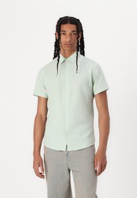 Camicetta verde chiaro con maniche corte e colletto button-down, caratterizzata da una vestibilità rilassata e una texture sottile, abbinata a pantaloni di denim chiaro.