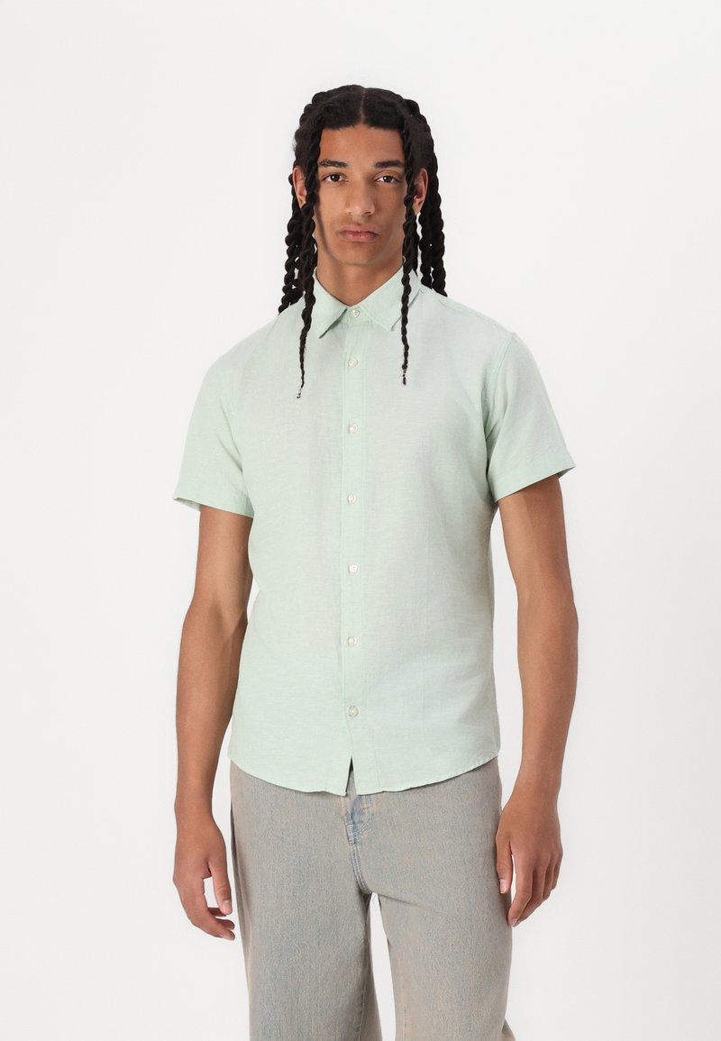 Camicetta verde chiaro con maniche corte e colletto button-down, caratterizzata da una vestibilità rilassata e una texture sottile, abbinata a pantaloni di denim chiaro.