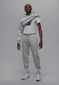 Grå sweatpants og matchende hættejakke, båret over en hvid t-shirt med et blåt grafik. Hvide sneakers med røde og sorte detaljer. Uformelt atletisk tøj.