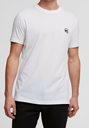 Mężczyzna ubrany w prosty biały t-shirt z krótkim rękawem, z małym czarno-białym logo na lewej piersi, w połączeniu z czarnymi spodniami.