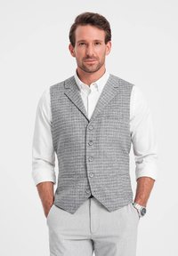 Chaleco a cuadros gris con cinco botones, llevado sobre una camisa blanca. El diseño presenta un corte entallado con un bolsillo en el lado izquierdo.