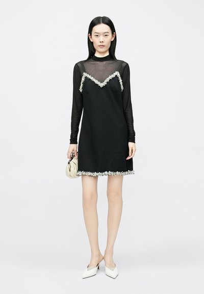 Melna kleita ar caurspīdīgām garām piedurknēm, ziedu rotājumi pie kakla izgriezuma un apakšmalas, kopā ar baltām smailām kurpēm un krēmkrāsas somiņu.