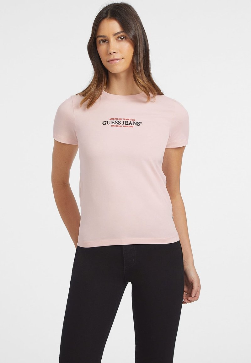 Camiseta ajustada de color rosa con mangas cortas que presenta un logo de "Guess Jeans" en negro y rojo en el frente. Textura suave, diseño minimalista.