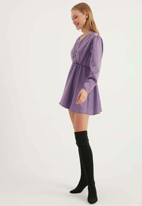Lila Minikleid mit langen Ärmeln, tailliertem Schnitt und V-Ausschnitt. Kombiniert mit knielangen schwarzen Stiefeln mit glatter Textur.