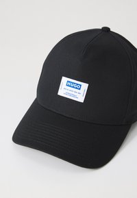Sort cap lavet af stof med en struktureret krone og buet skygge. Har et rektangulært hvidt label med blå tekst, der står "HUGO."