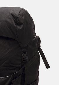 Czarny plecak z tkaniny z zaokrąglonym dnem, regulowanymi szelkami, plastikowym zapięciem i bocznymi kieszeniami. Posiada teksturowane wykończenie.