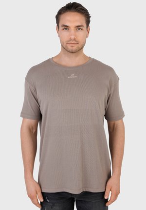 Mężczyzna o krótkich ciemnych włosach, ubrany w taupeową koszulkę z okrągłym dekoltem z małym białym logo na środku klatki piersiowej, w połączeniu z czarnymi jeansami.