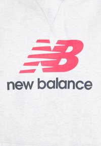 Rotes stilisiertes "NB"-Logo über schwarzem "new balance"-Text auf einem hellgrauen Stoffhintergrund.