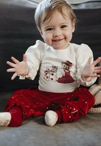 Maglietta bianca a maniche lunghe con maniche a sbuffo che presenta una grafica di gatto e ragazza, abbinata a pantaloni di velluto rossi decorati con piccoli cuori dorati.