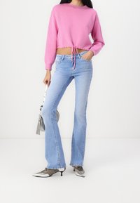 Rosa kortad tröja med långa ärmar och dragsko i fållen, ihop med blå utsvängda jeans och skor med silverdetaljer. Rund metallhandväska.