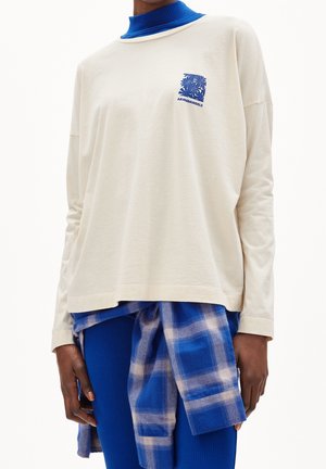 Osoba ubrana w kremową koszulę z długim rękawem z niebieskim kwiatowym logo, królewsko niebieski golf, niebieskie prążkowane spodnie i niebieską kratkowaną koszulę zawiązaną w pasie.