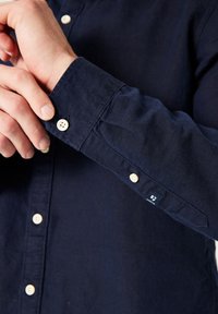 Camisa de botones azul marino con tejido texturizado, botones blancos y una etiqueta de la marca en la zona del puño. Corte slim fit con mangas largas.