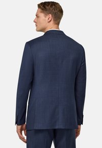 Veste de costume ajustée bleu maritime, tissu texturé, fente centrale, revers crantés et poignets boutonnés. Détails de couture visibles au niveau des coutures.