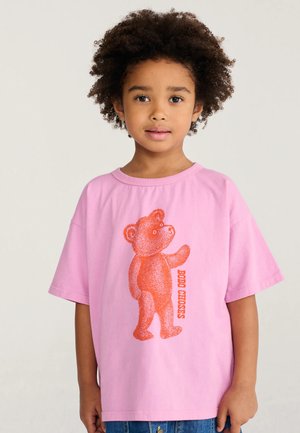 Niño pequeño con cabello rizado que lleva una camiseta rosa con un gráfico de un osito naranja y el texto "BOBO CHOSES" en la parte delantera.