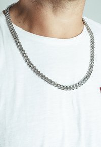 Collana in metallo argentato a catena, con un design a link spesso. Indossata su una semplice maglietta bianca, evidenziando una texture liscia e lucida.