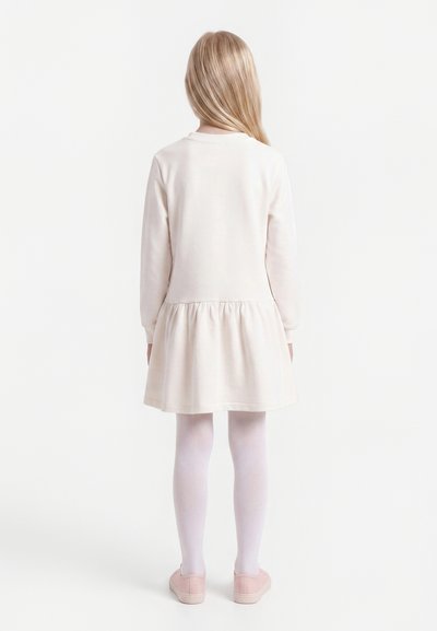 Jeune fille aux longs cheveux blonds portant une robe rose pâle à manches longues, des collants blancs et des chaussures roses, debout de dos.