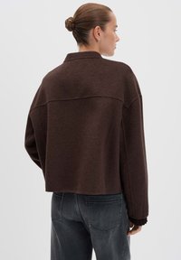 Vrouw met haar in een knot, gekleed in een donkerbruin kort jasje en vervaagde zwarte jeans, met de rug naar de camera tegen een effen achtergrond.