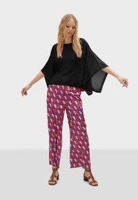 Černý volný top s širokými rukávy, doplněný jasně vzorovanými růžovými kalhotami se geometrickými tvary. Bronzové sandály na řemínky dotvářejí vzhled.