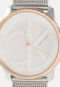 Montre ronde avec un bracelet en maille argentée, un cadran blanc texturé, des accents dorés et un cadran minimaliste avec un logo et une aiguille des heures simple.