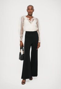 Blusa blanca transparente con mangas abullonadas y cuello atado; pantalones anchos negros, bolso negro y tacones con tiras completan el conjunto.