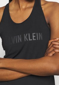 Mustat hihaton paita, jossa on pyöreä pääntie, ja rinnassa kohotettua "CALVIN KLEIN" -tekstiä. Sileä kangas, tiukka malli.
