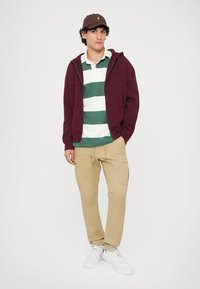 Bordeaux zip-up hoodie, grøn og hvid stribet polo, lysebrune cargo bukser og hvide sneakers. Inkluderer en brun cap med et broderet logo.