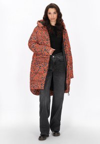 Vadderad jacka i livlig orange med blå leopardmönster, med huva, främre blixtlås och sidofickor. Kombinerad med svarta vida jeans.