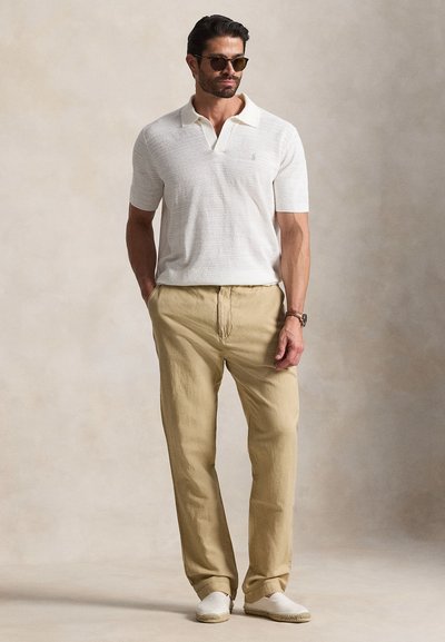 Άνδρας με λευκό κοντομάνικο πόλο, μπεζ παντελόνι, λευκά παπούτσια χωρίς κορδόνια, γυαλιά ηλίου και ρολόι, στέκεται μπροστά σε απλό ουδέτερο φόντο.