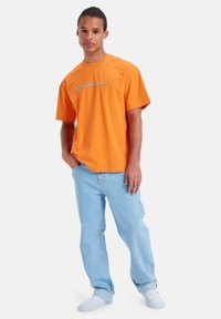 Oranžové bavlněné tričko s volným střihem, kulatým výstřihem a logem "Quiksilver" v kontrastní barvě. Kombinováno se světle modrými džínami.