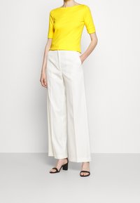 Gulfärgad tajt topp med korta ärmar, matchad med vita vida byxor. Looken kompletteras av svarta klacksandaler.